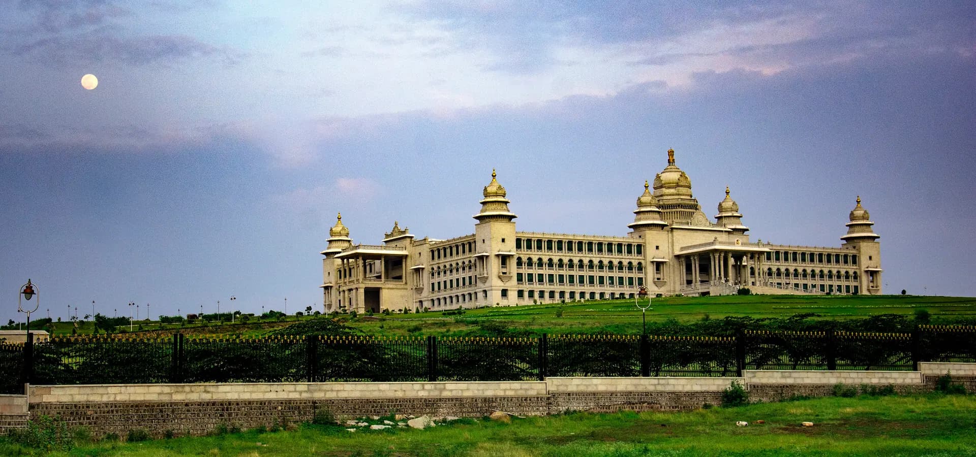Das weiße Suvarna Vidhana Soudha mit Kuppel auf einem grünen Hügel in der Dämmerung, der Mond steigt an einem weichen violetten Himmel auf