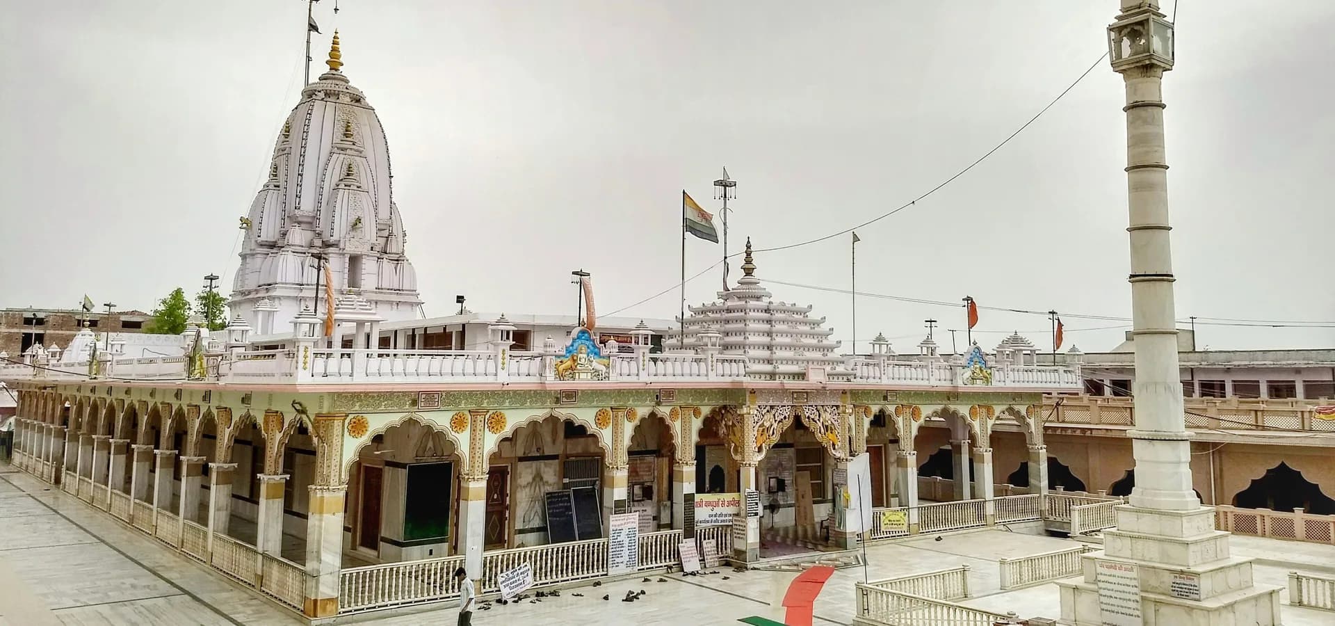 White marble spire and tall pillar of the Tijara Jain temple above an arcaded courtyard under a pale sky