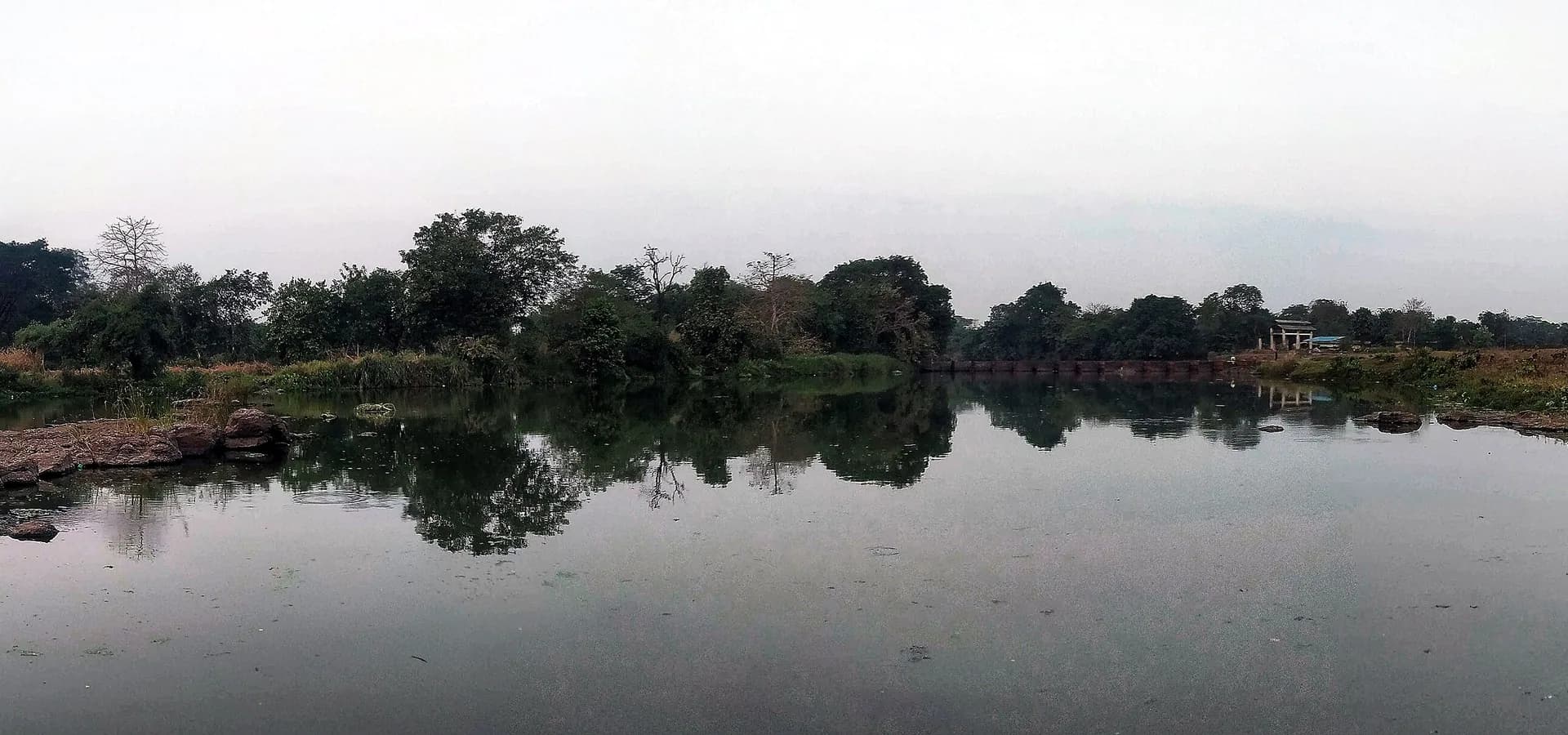 Vista amplia de árboles verdes y rocas reflejados en un río tranquilo bajo un suave cielo gris cerca de Bhiwandi