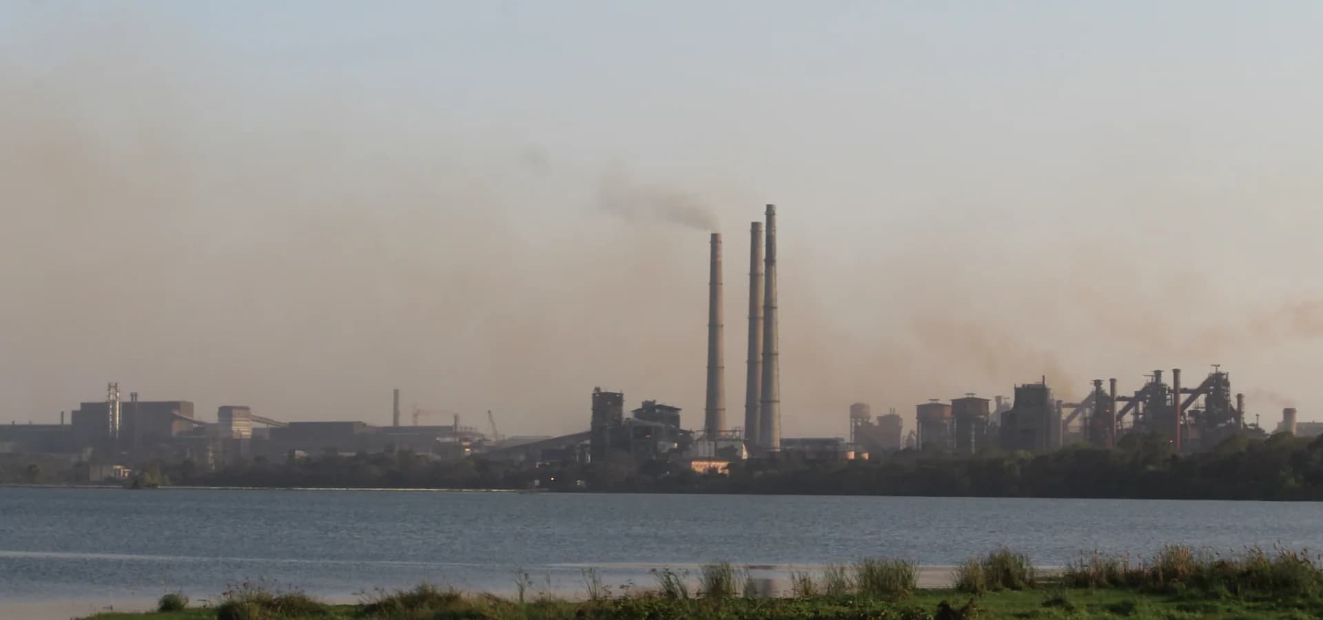 Tall chimneys and blast furnaces of the Bokaro Steel Plant on the far shore of a calm lake with green banks