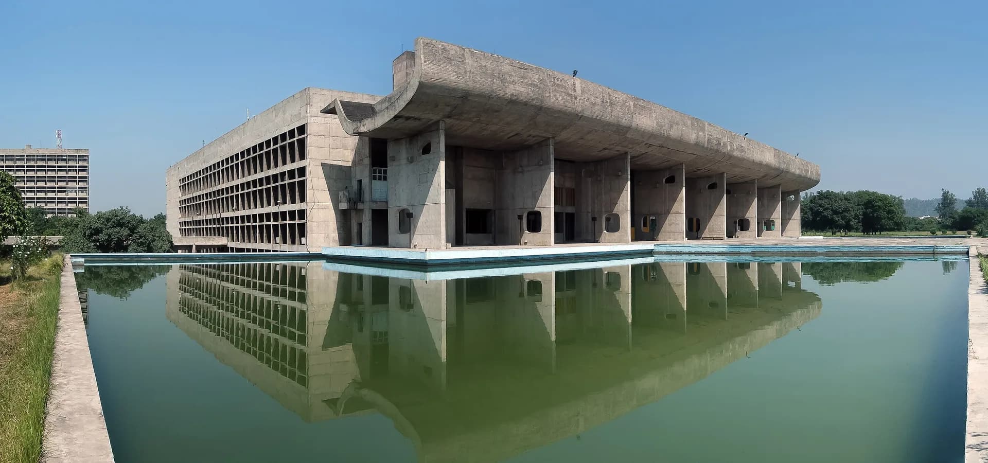 Der Palace of Assembly aus Beton mit gewölbtem Dach, gespiegelt in einem langen grünen Becken unter blauem Himmel