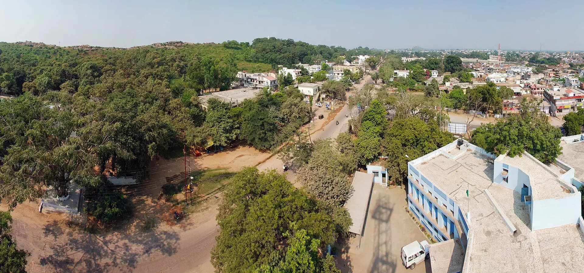 Amplia vista aérea de Chhatarpur, con una colina boscosa verde a la izquierda y techos bajos entre árboles bajo un cielo despejado