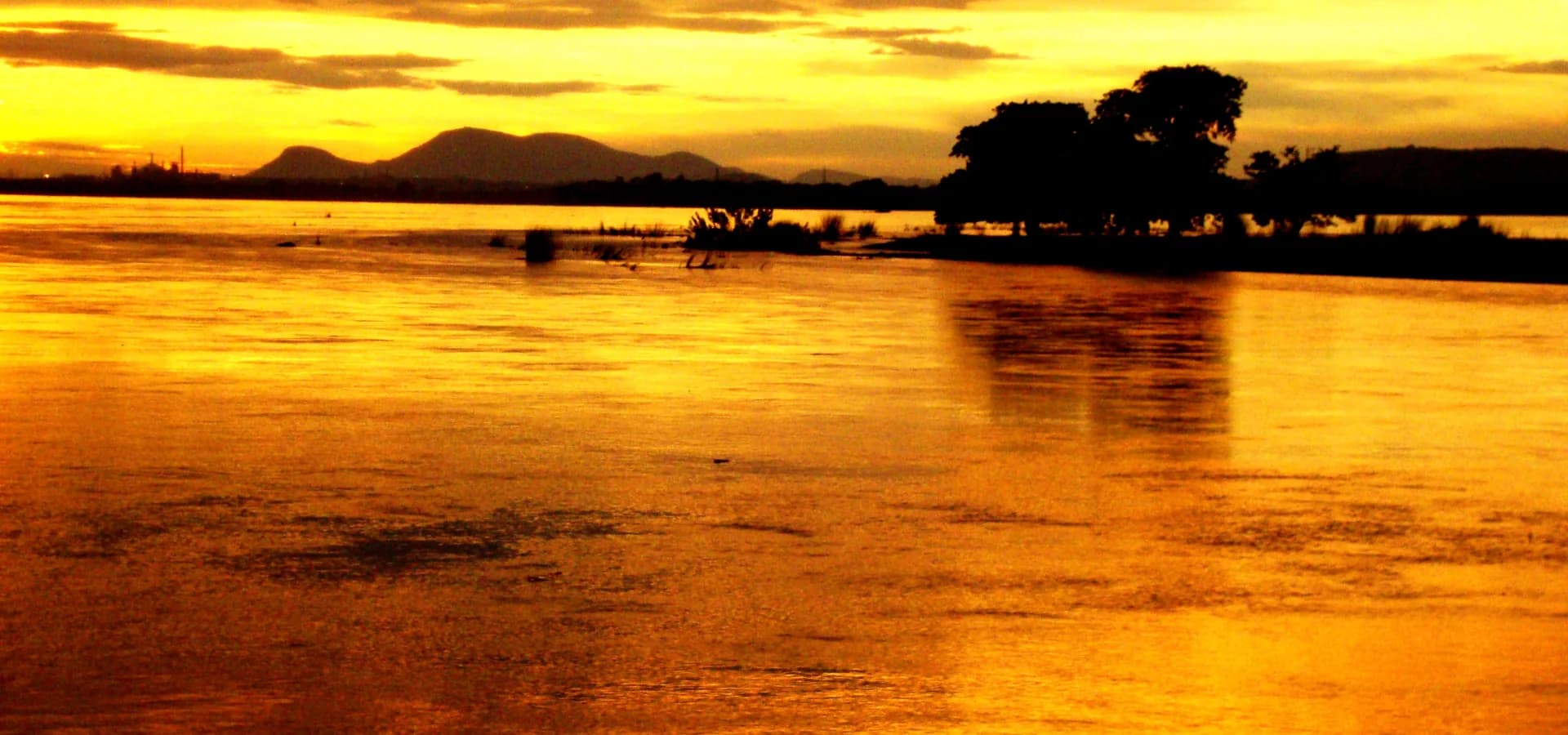 El ancho río Mahanadi en Chahata, Cuttack, dorado al atardecer, con colinas bajas y árboles en la otra orilla