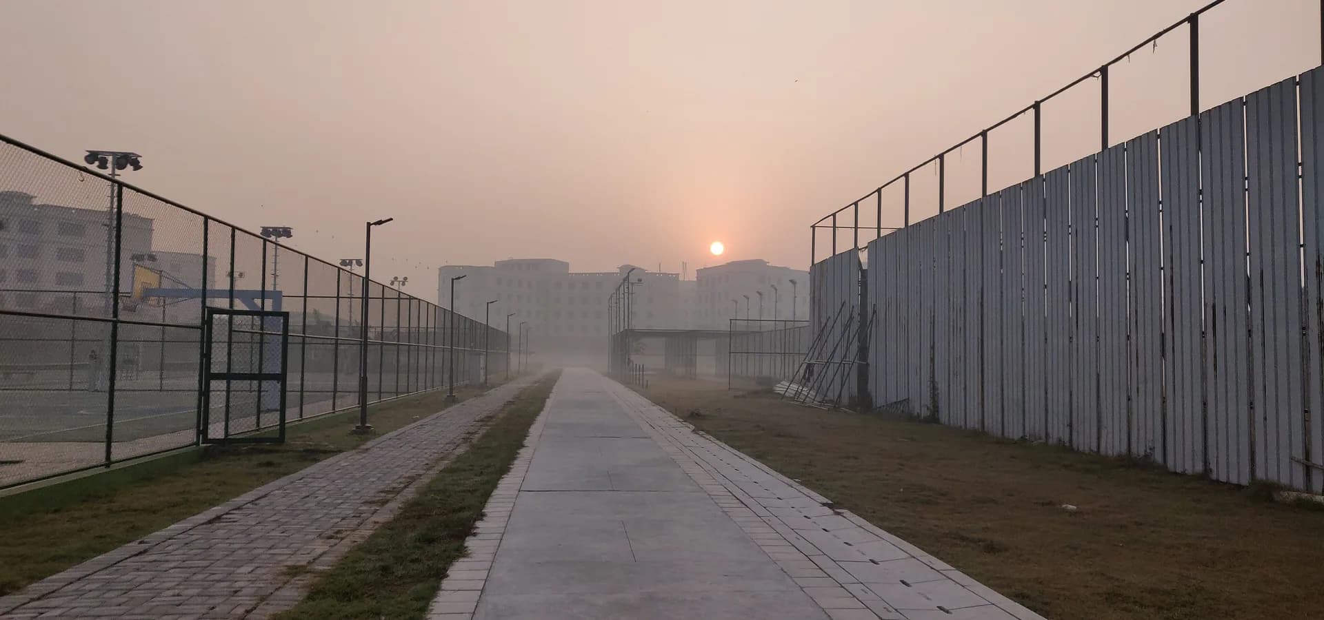 Un sol naranja pálido sale entre la neblina invernal sobre un largo sendero pavimentado y edificios del campus cerca de Dadri