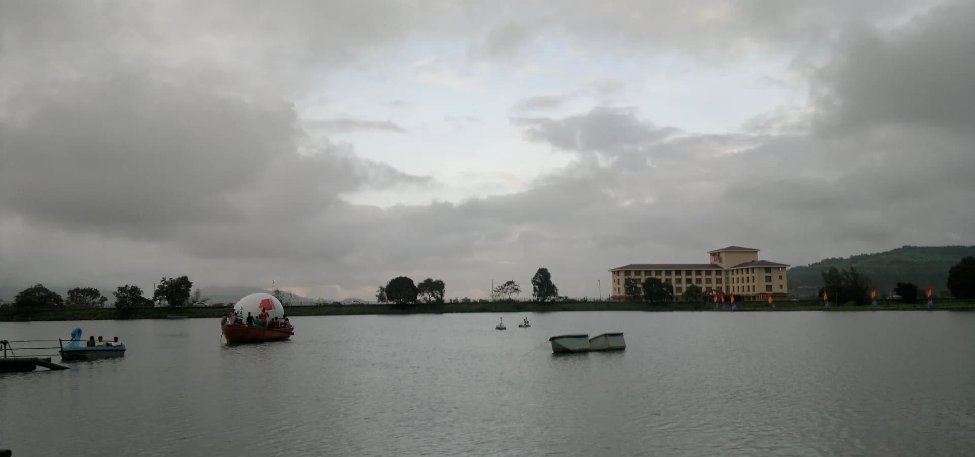 Vista ampla do Lago Saputara com pedalinhos, um hotel à beira do lago e colinas verdes sob um céu nublado de monção