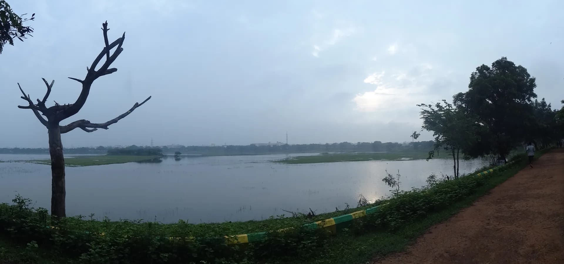 Kunduvada Kere en Davanagere, un amplio lago tranquilo bajo un cielo pálido con un sendero junto a la orilla y árboles