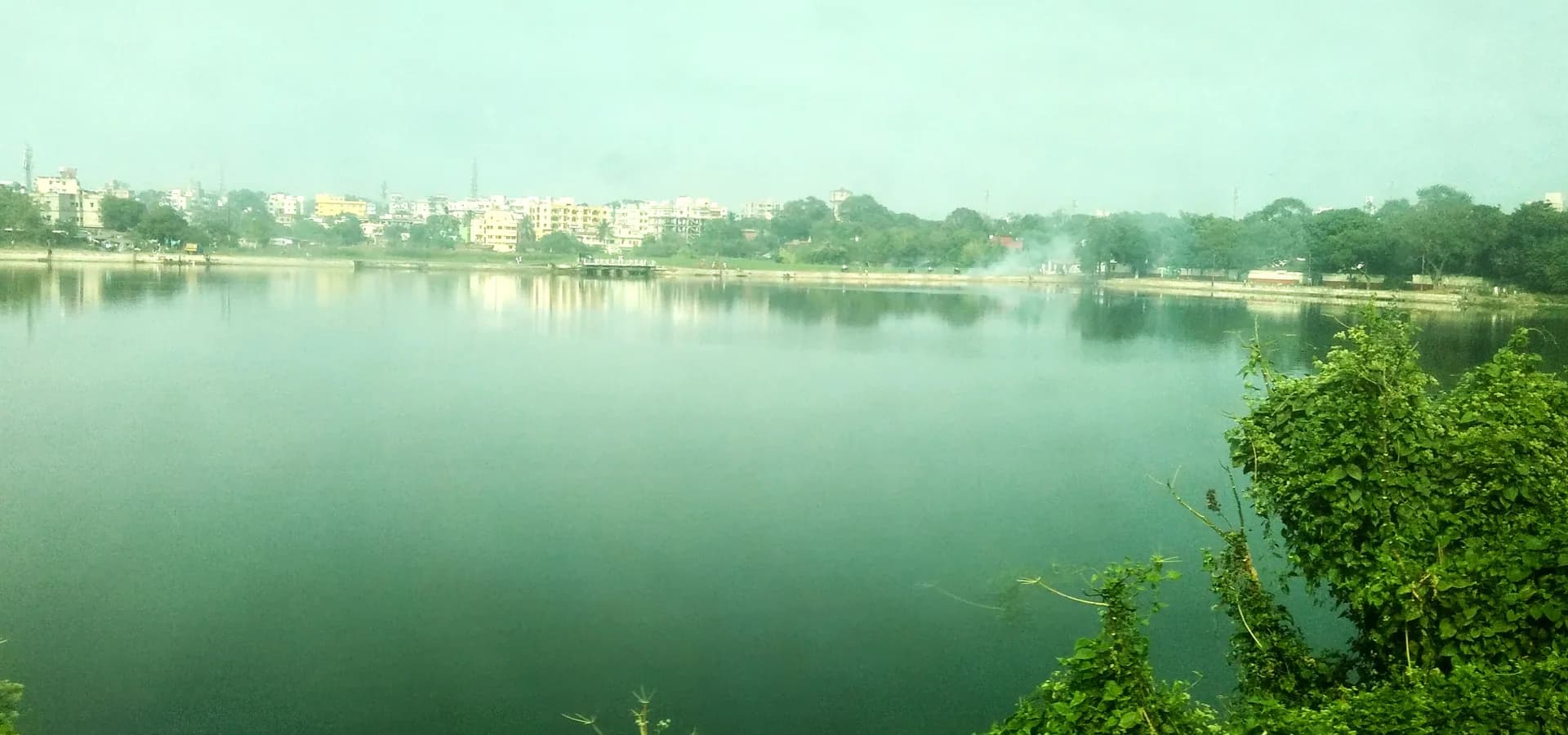 A água verde e calma do Loco Tank em Dhanbad, com prédios de apartamentos e árvores ao longo da margem oposta