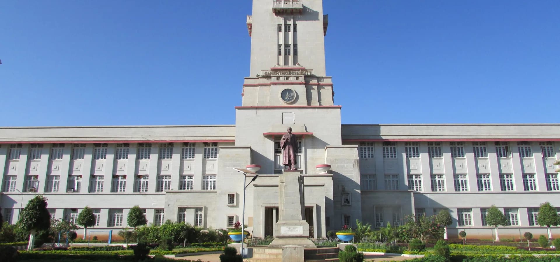 Prédio branco da Karnatak University com uma torre de relógio de cúpula rosa sobre jardins aparados e um céu azul