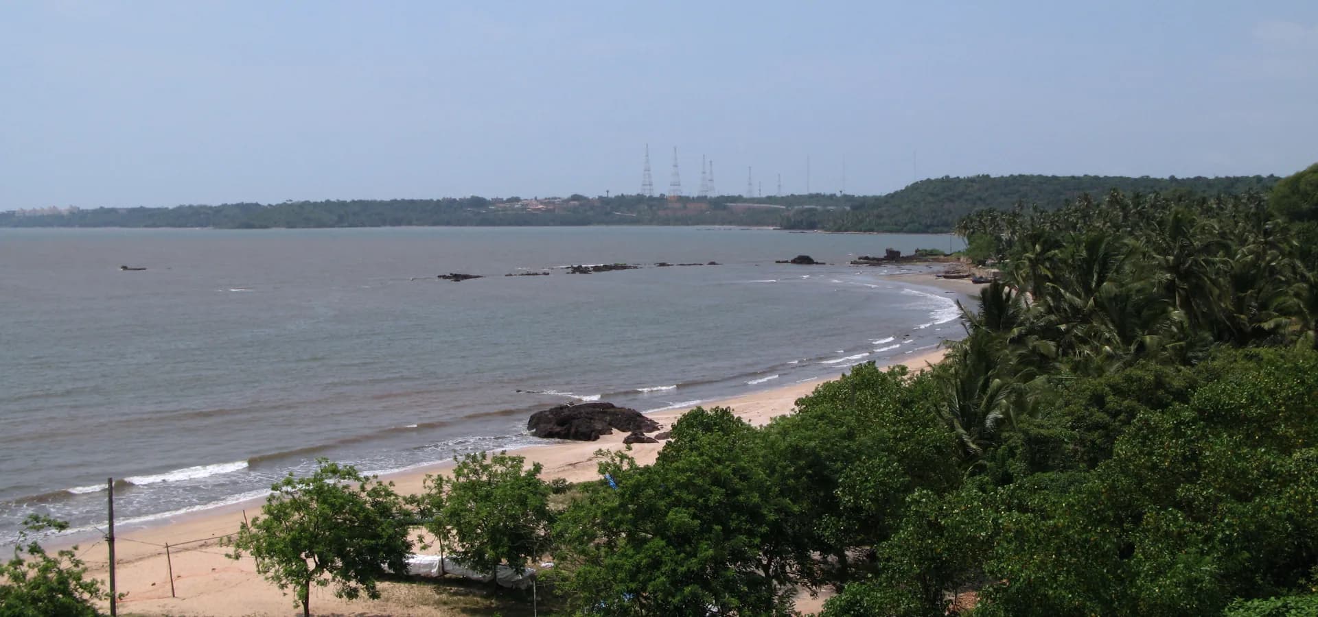 Sandiger Strand von Siridao in einer Kurve entlang der ruhigen Zuari-Mündung, mit grünen Bäumen und Palmen im Vordergrund