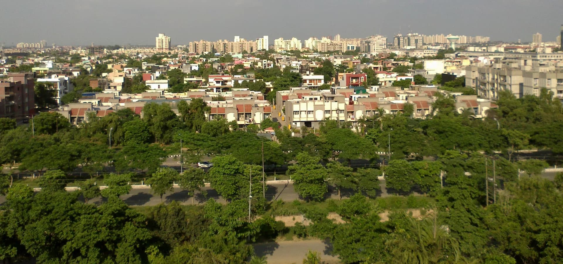 Greater Noida vista do alto, com ruas arborizadas e casas baixas dando lugar a torres de apartamentos ao longe sob um céu azul