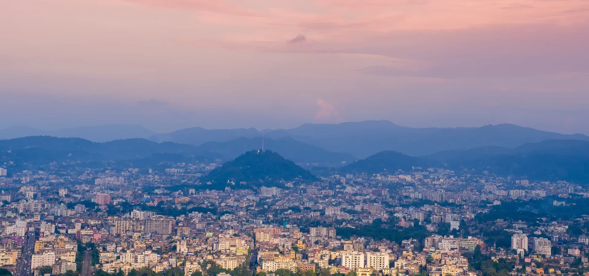 नीलाचल पहाड़ी से गुलाबी शाम के आसमान के नीचे जंगली पहाड़ियों की ओर फैला गुवाहाटी