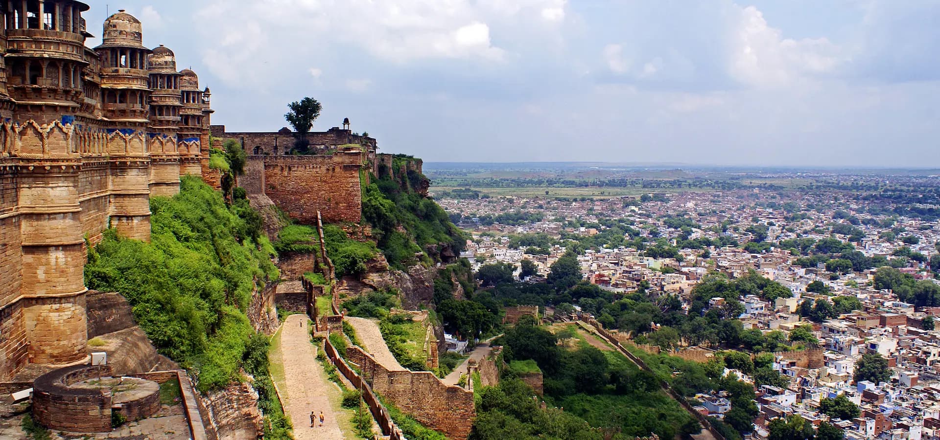 Kuppeltürme und Festungsmauern des Gwalior Fort auf einem grünen Hügel über der Stadt unter bewölktem Himmel
