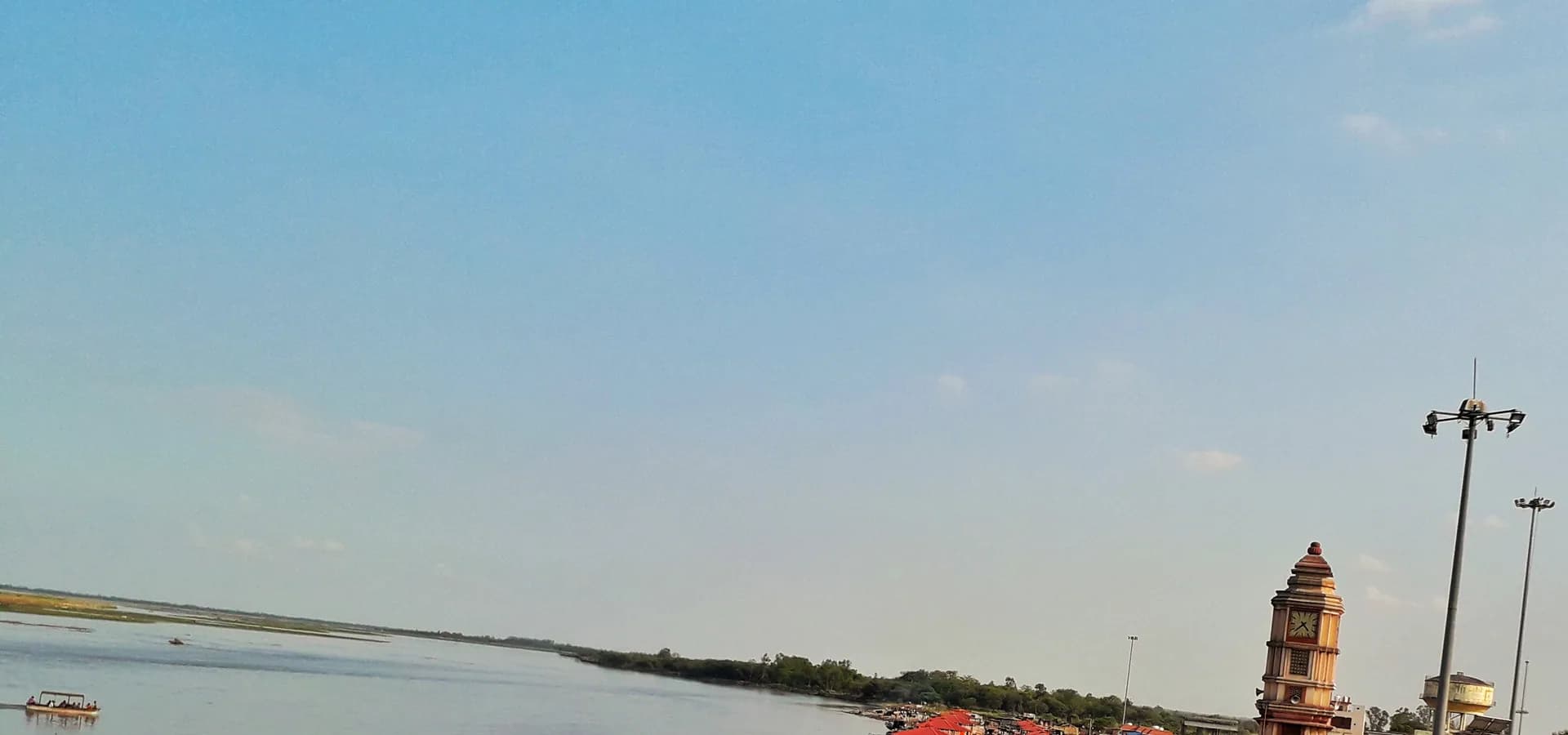 Weiter Blick auf den Ganga bei Brijghat unter klarem Himmel, mit Uhrturm und rotbedachten Ghats am rechten Ufer