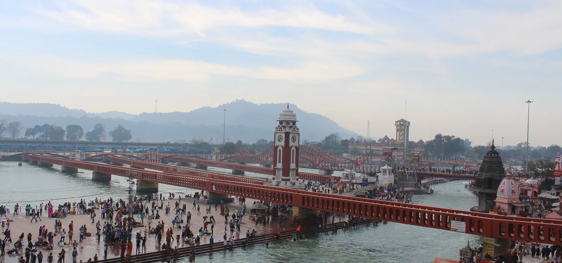 Footbridges cross the Ganges at Har Ki Pauri in Haridwar, with the clock tower, crowded ghats and hazy hills behind