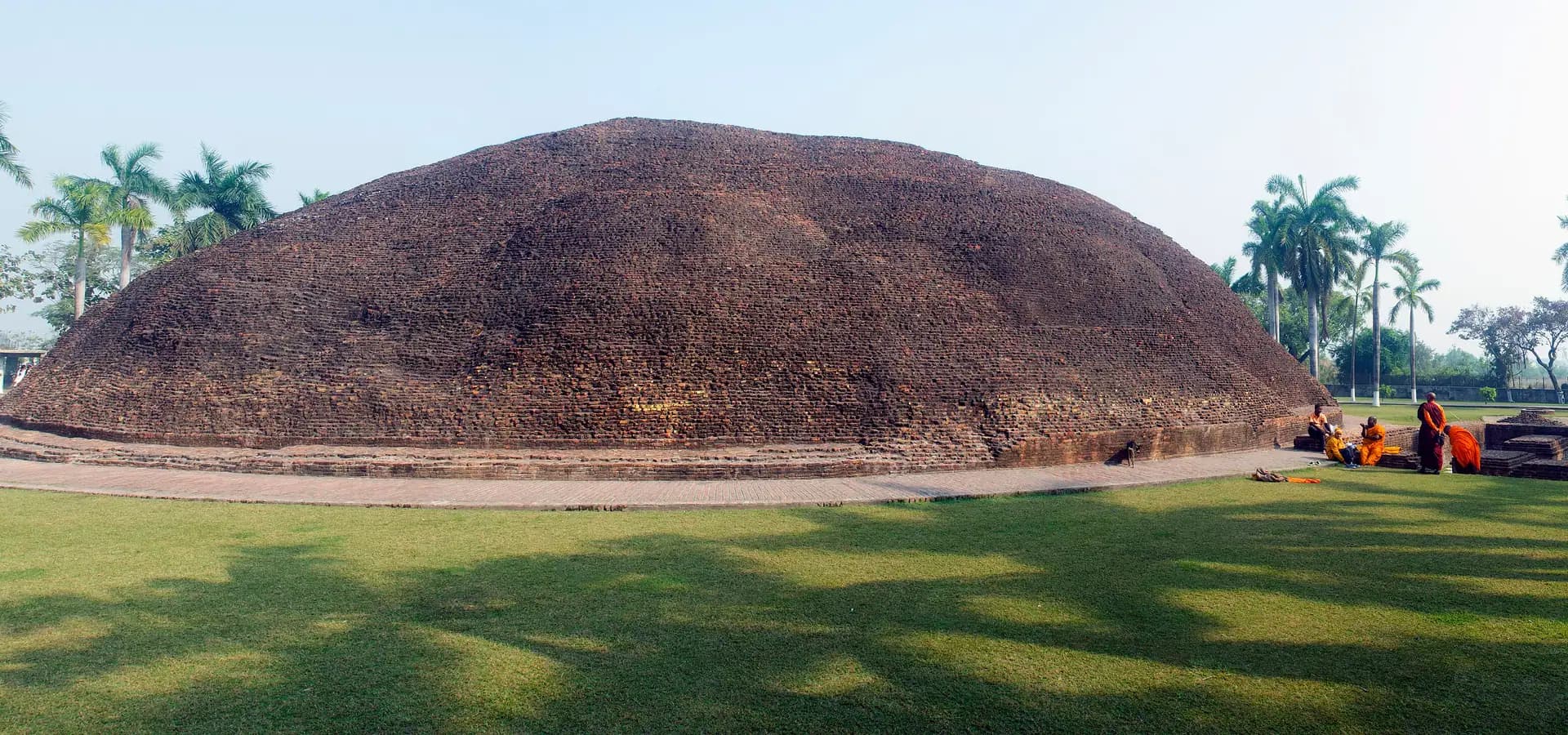 Der riesige runde Backsteinstupa Ramabhar auf grünem Rasen unter blassem Himmel, umgeben von Palmen in Kushinagar