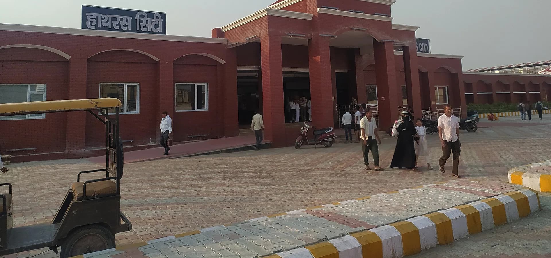 Red-brick facade of Hathras City railway station with a gabled entrance, paved forecourt and a wide pale sky