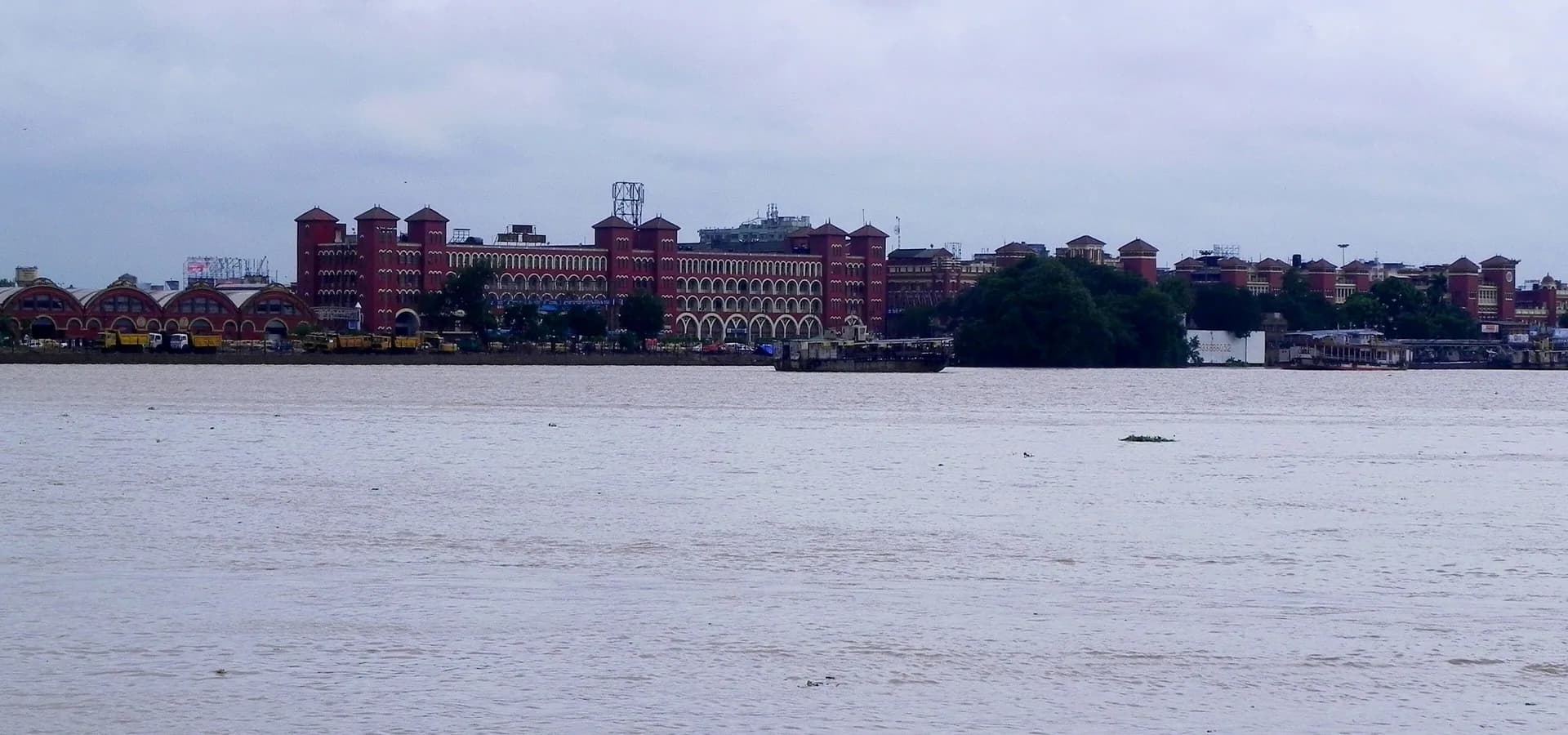 Das lange rote Backsteingebäude des Bahnhofs Howrah mit seinen Türmen, über den breiten Hooghly unter grauen Wolken gesehen