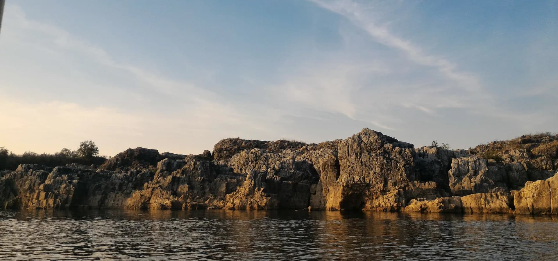 Penhascos baixos de mármore margeiam o calmo Rio Narmada em Bhedaghat sob um céu claro de fim de tarde com nuvens finas