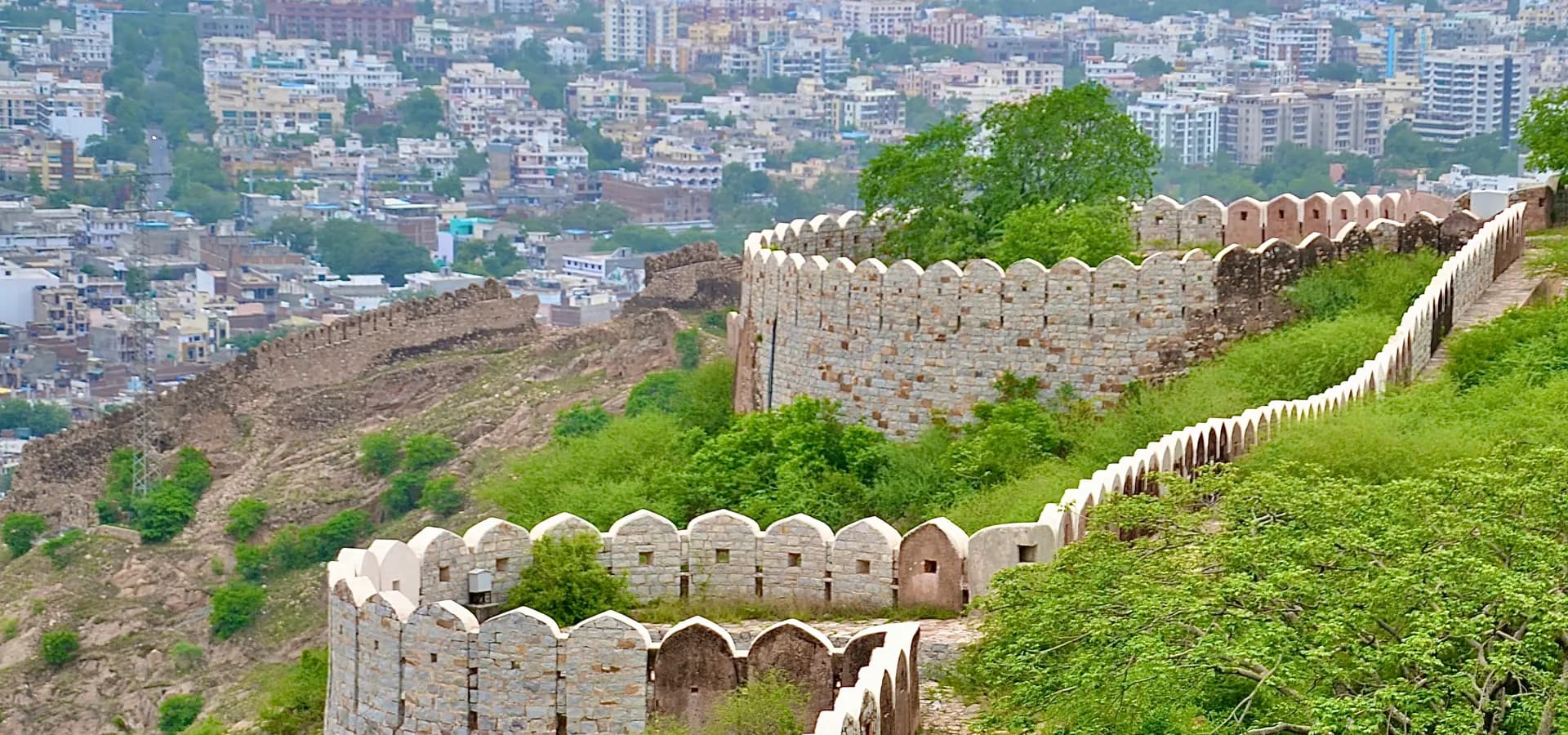 Zinnenbewehrte Mauern des Nahargarh Fort schwingen sich einen grünen Hang entlang, darunter breitet sich Jaipur aus