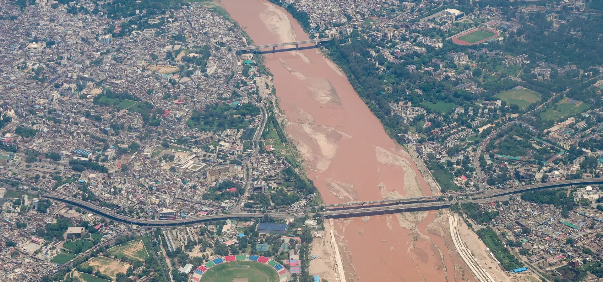 Vista aérea de Jammu con el ancho río Tawi, dos puentes y un estadio de críquet entre barrios densos