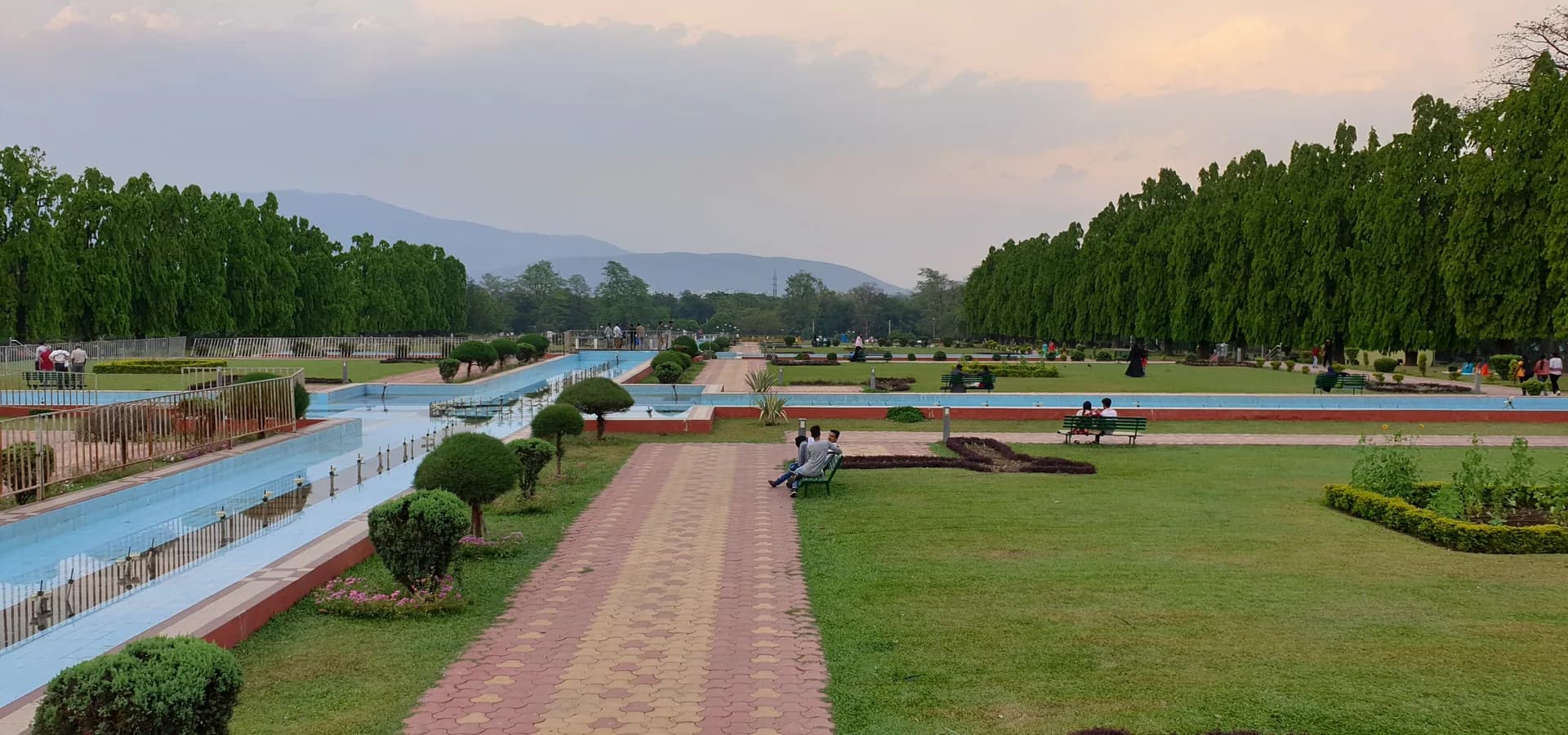 Weite Rasenflächen und lange Wasserkanäle im Jubilee Park, von hohen Bäumen gesäumt, mit blauen Dalma Hills am Horizont