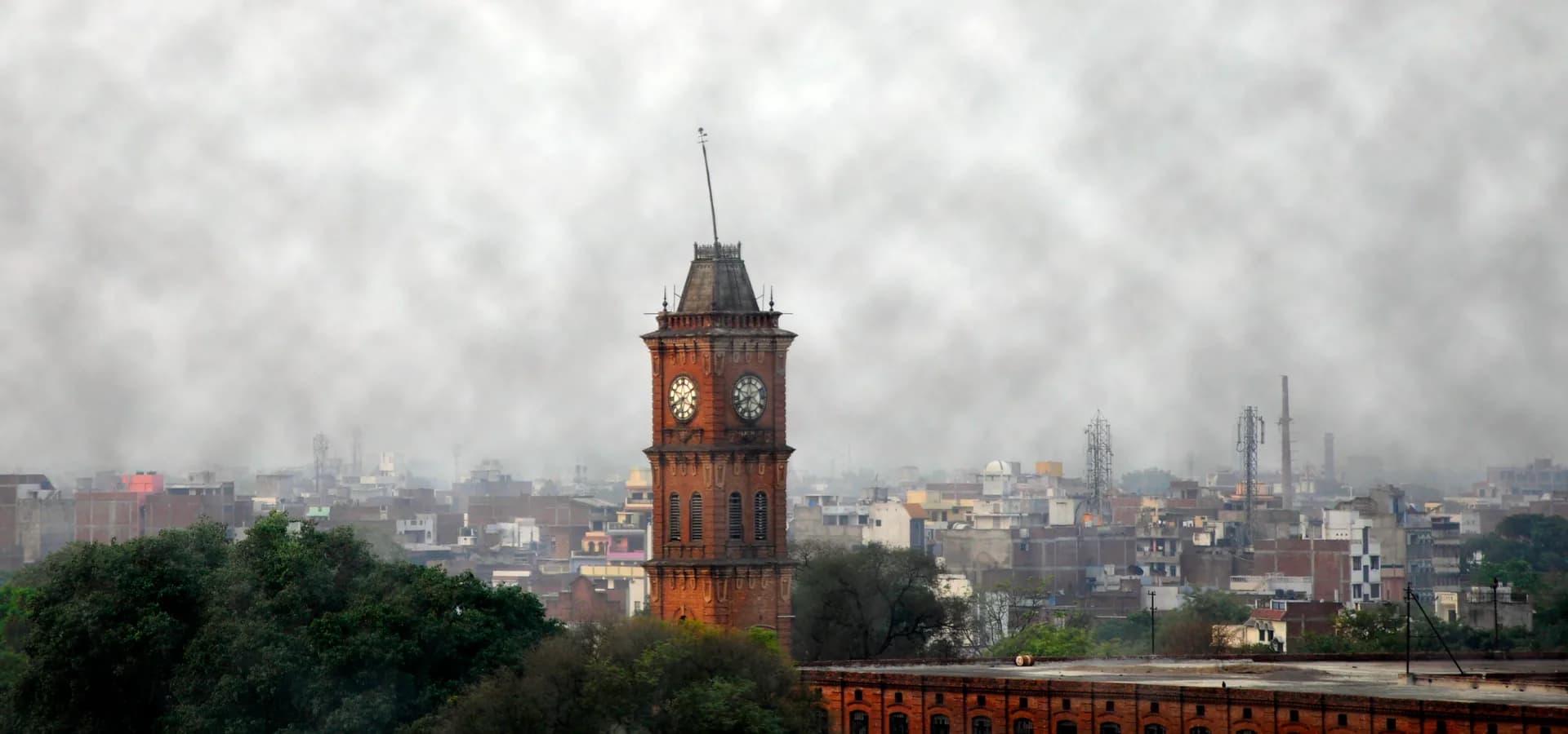 A torre de relógio de tijolos vermelhos Lal Imli se erguendo sobre árvores e telhados de Kanpur sob um céu nublado e pálido