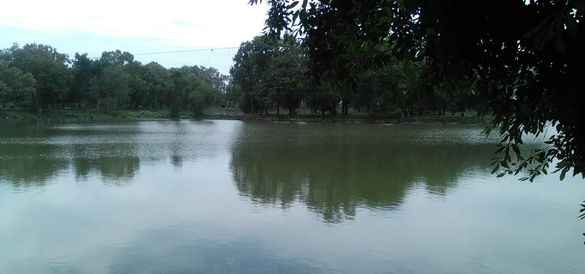 Stilles Wasser des Karna Lake, das die Bäume am anderen Ufer und einen weichen Wolkenhimmel nahe Karnal spiegelt