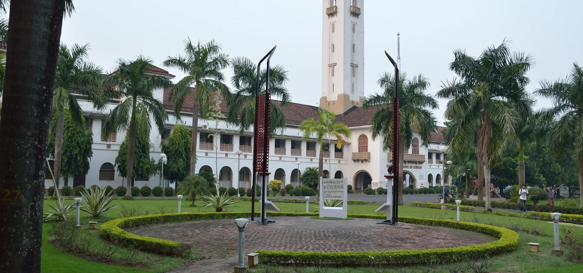 IIT खड़गपुर परिसर में ताड़ के पेड़ों और गोल लॉन के पीछे ऊंची मीनार वाला सफेद हिजली शहीद भवन