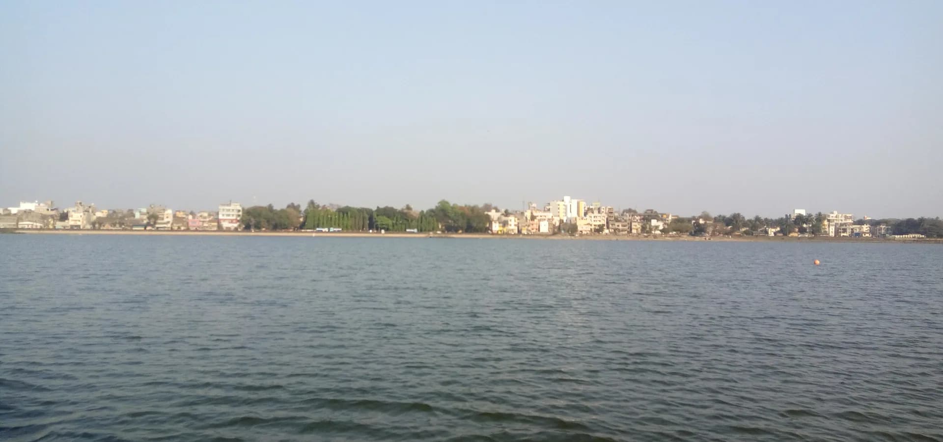 Aguas tranquilas del lago Rankala hasta una baja línea de edificios y árboles de Kolhapur bajo un cielo despejado