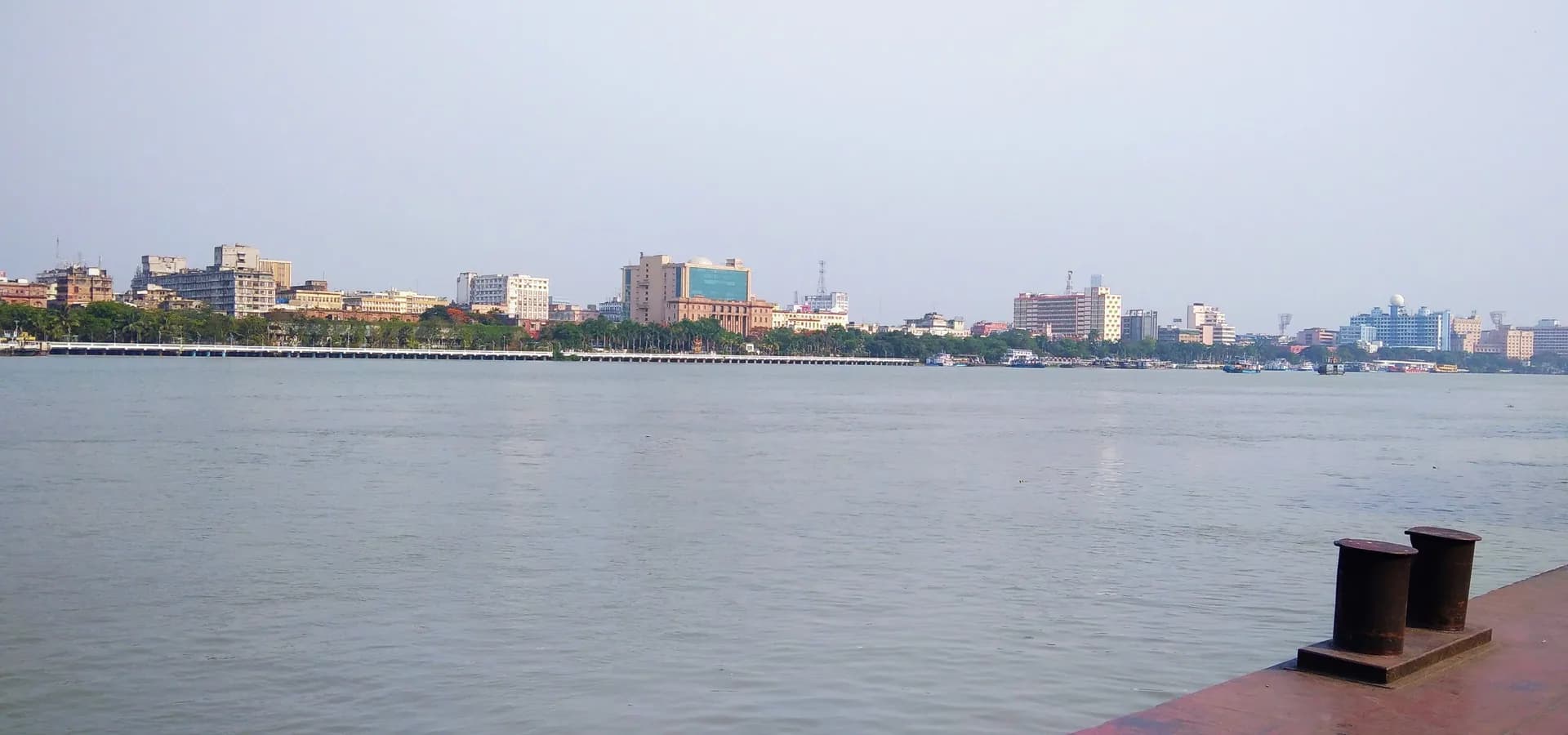 Kolkata riverfront buildings and palm-lined promenade seen across the calm Hooghly River on a hazy day