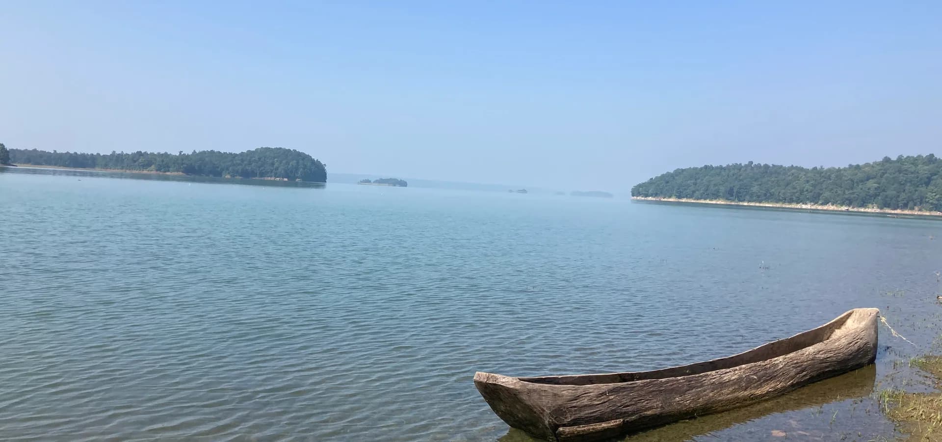 Ruhiger blauer Stausee mit bewaldeten Inseln bei Korba, ein hölzerner Einbaum liegt im Flachwasser