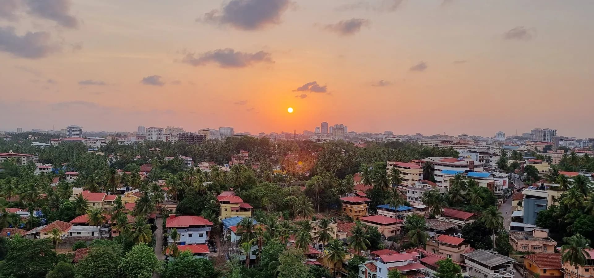 Sonnenuntergang über Mangaluru, vorn rote Ziegeldächer und Kokospalmen, am Horizont Stadttürme