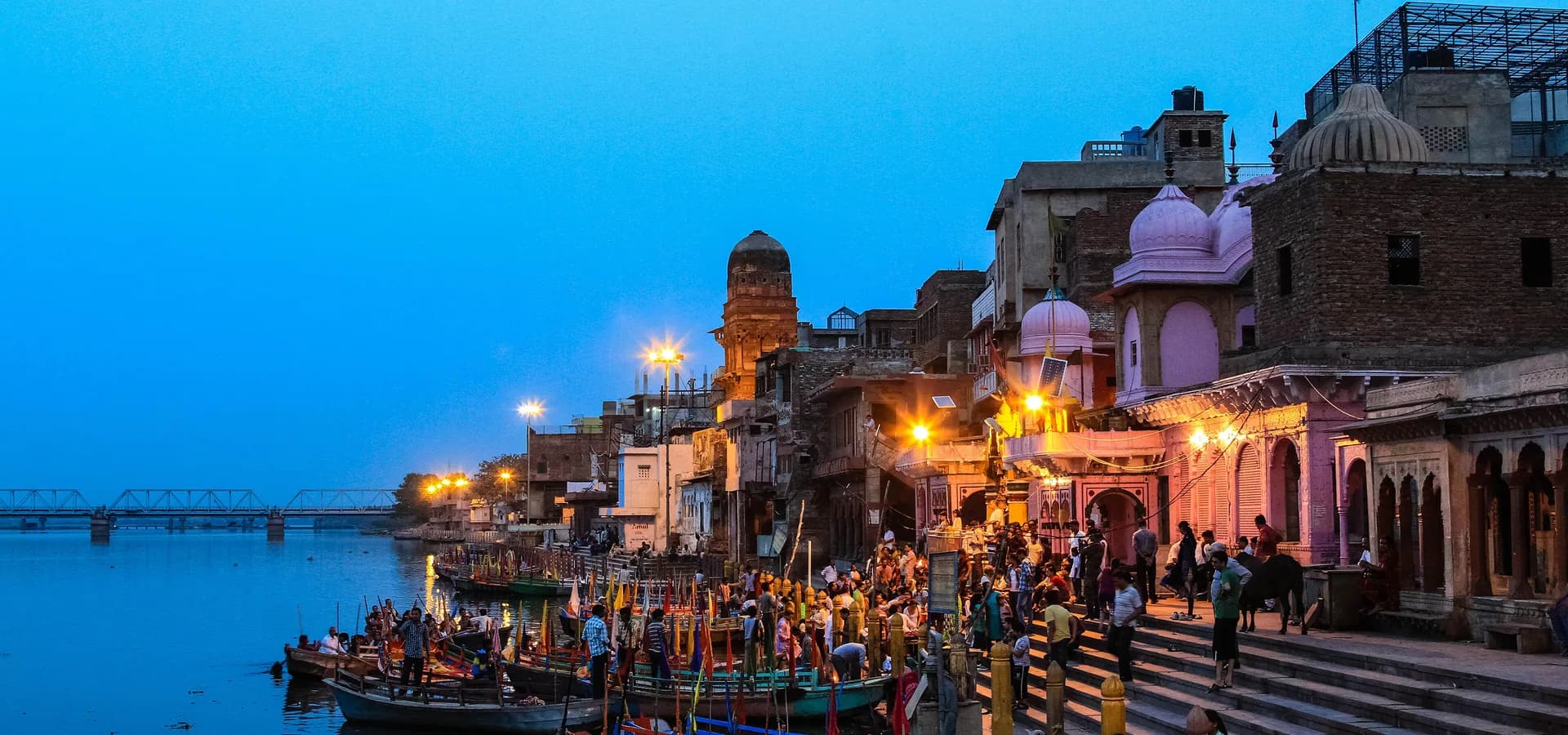 शाम के समय विश्राम घाट, यमुना में बँधी लकड़ी की नावें, ऊपर रोशन सीढ़ियाँ और गुलाबी गुंबद वाले मंदिर