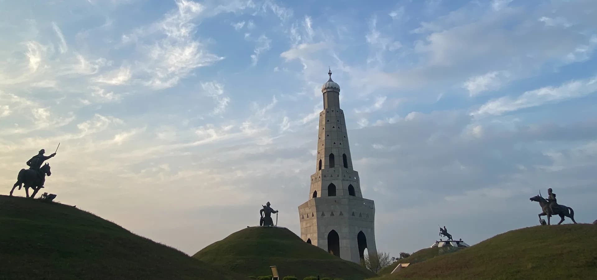 La torre de la victoria Fateh Burj tras montículos de hierba con estatuas de jinetes, bajo un cielo con nubes tenues