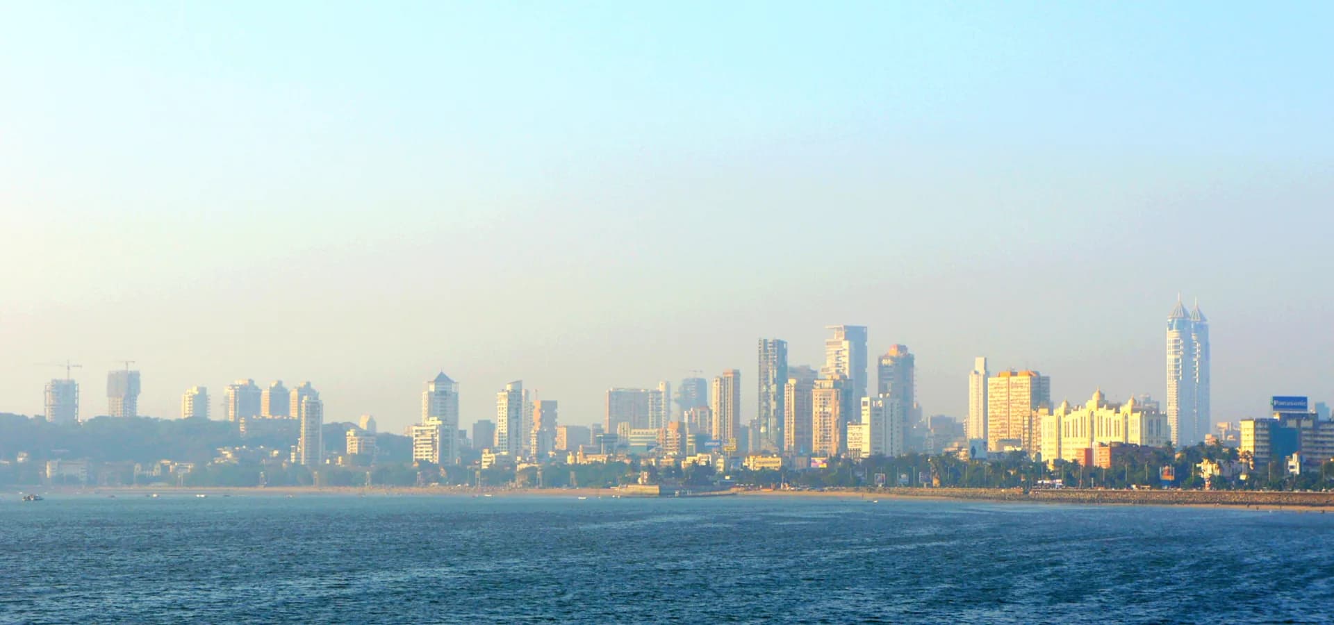 Hochhäuser von Mumbai am Ufer der Back Bay unter blassem Morgenhimmel, davor ruhiges blaues Wasser