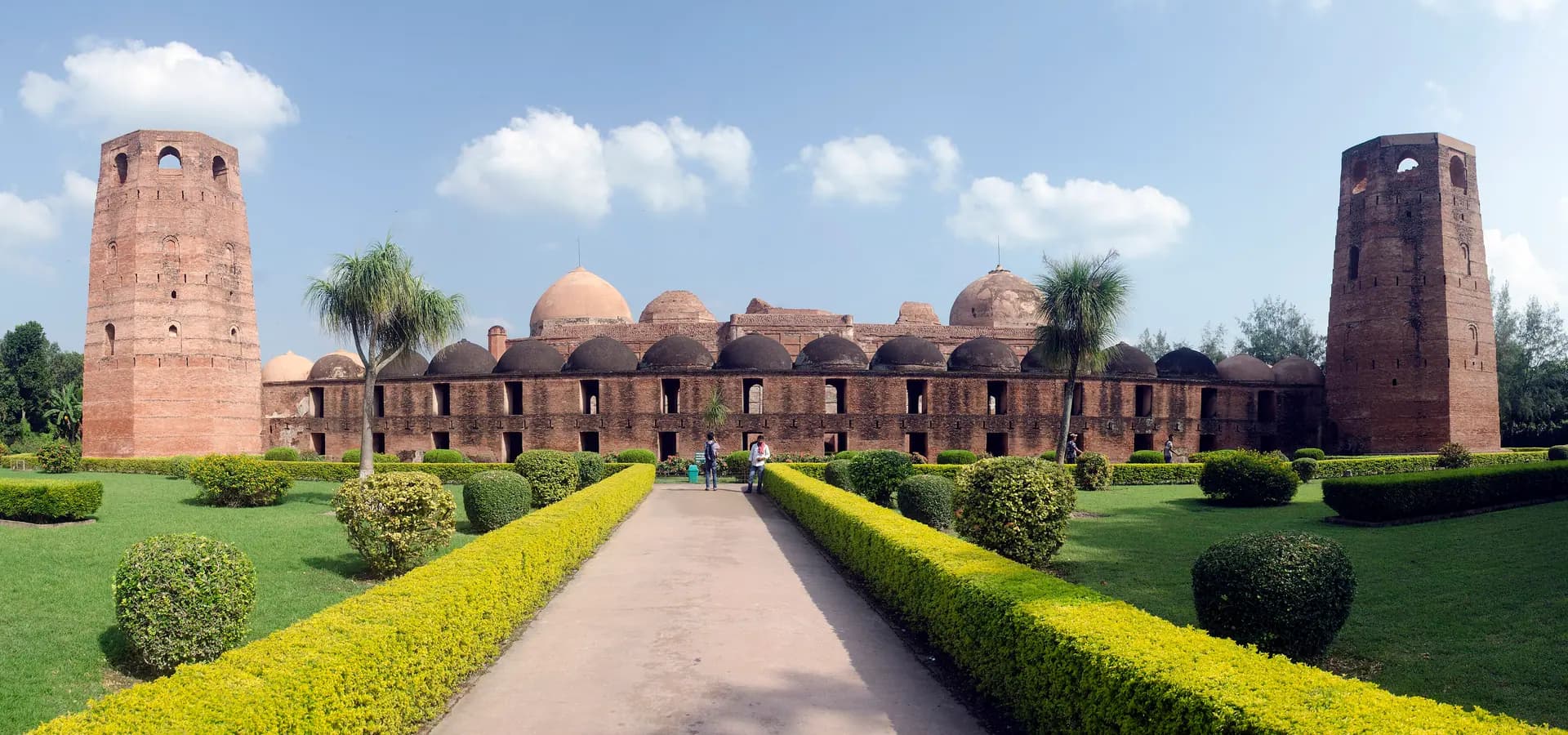 Vista ampla da Katra Masjid de tijolo em Murshidabad, duas altas torres de canto emoldurando fileiras de cúpulas sob céu azul