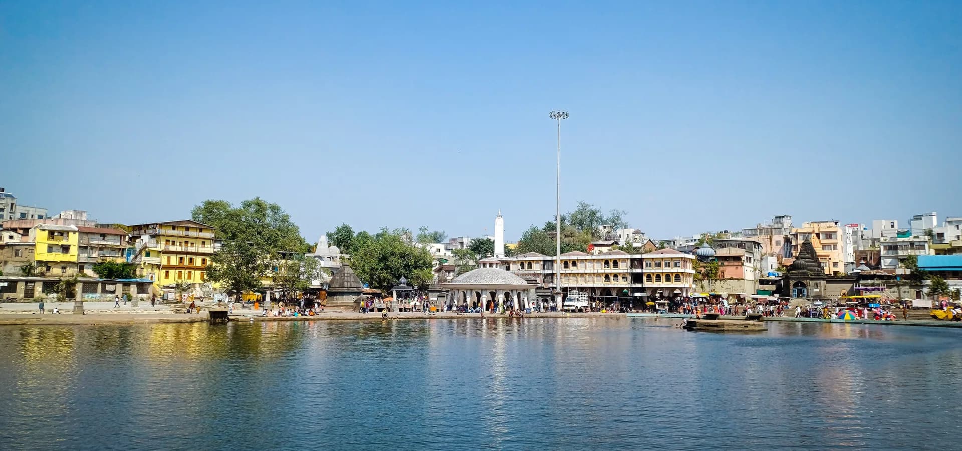 Ruhiges Godavari-Wasser am Gandhi Talav spiegelt die Ghats, kleinen Tempel und Häuser der Altstadt von Nashik unter blauem Himmel