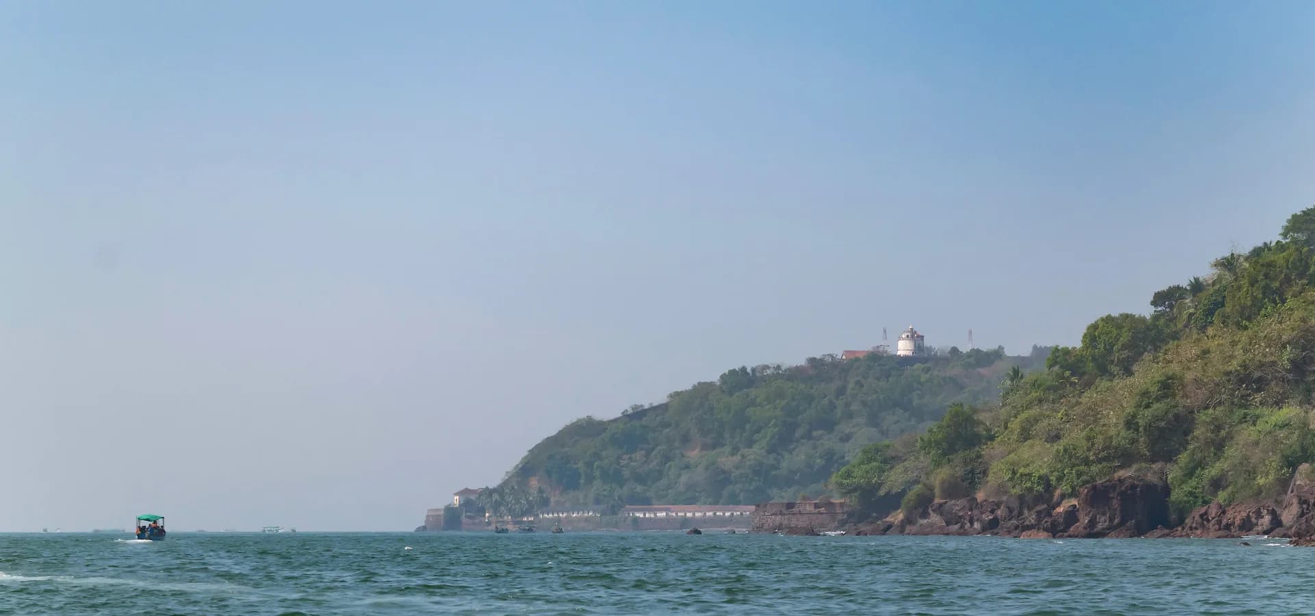 Grüne Landzunge mit den Mauern von Fort Aguada und einem weißen Leuchtturm über dem Arabischen Meer, vom Boot aus gesehen