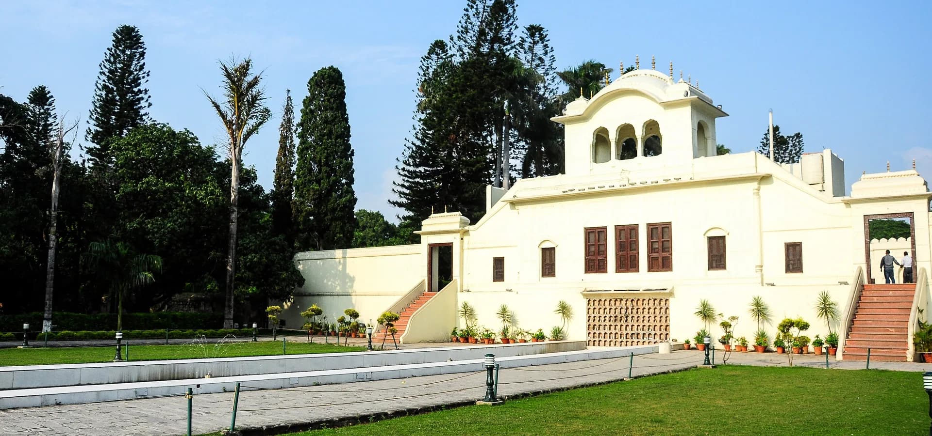 Palácio branco de estilo mogol com pavilhão de cúpula ao lado de pinheiros altos e um gramado verde nos Jardins de Pinjore