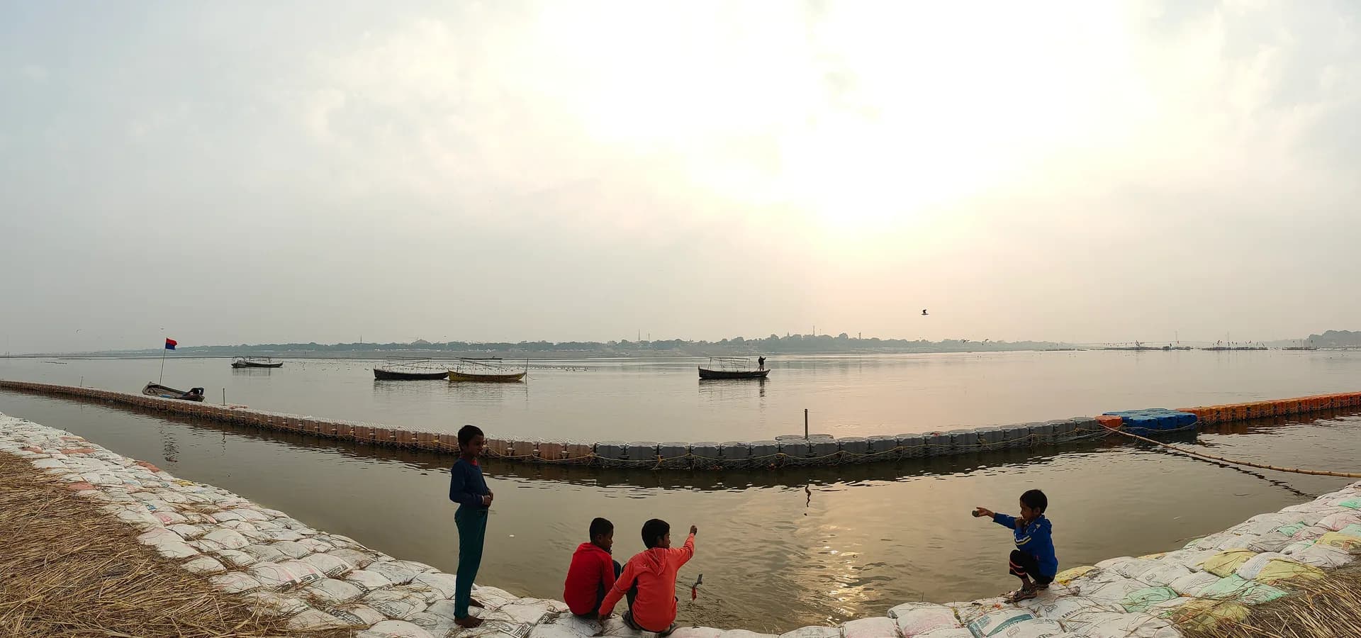 Wide hazy view over the Triveni Sangam in Prayagraj, rowboats resting on calm water near a sandbagged bank