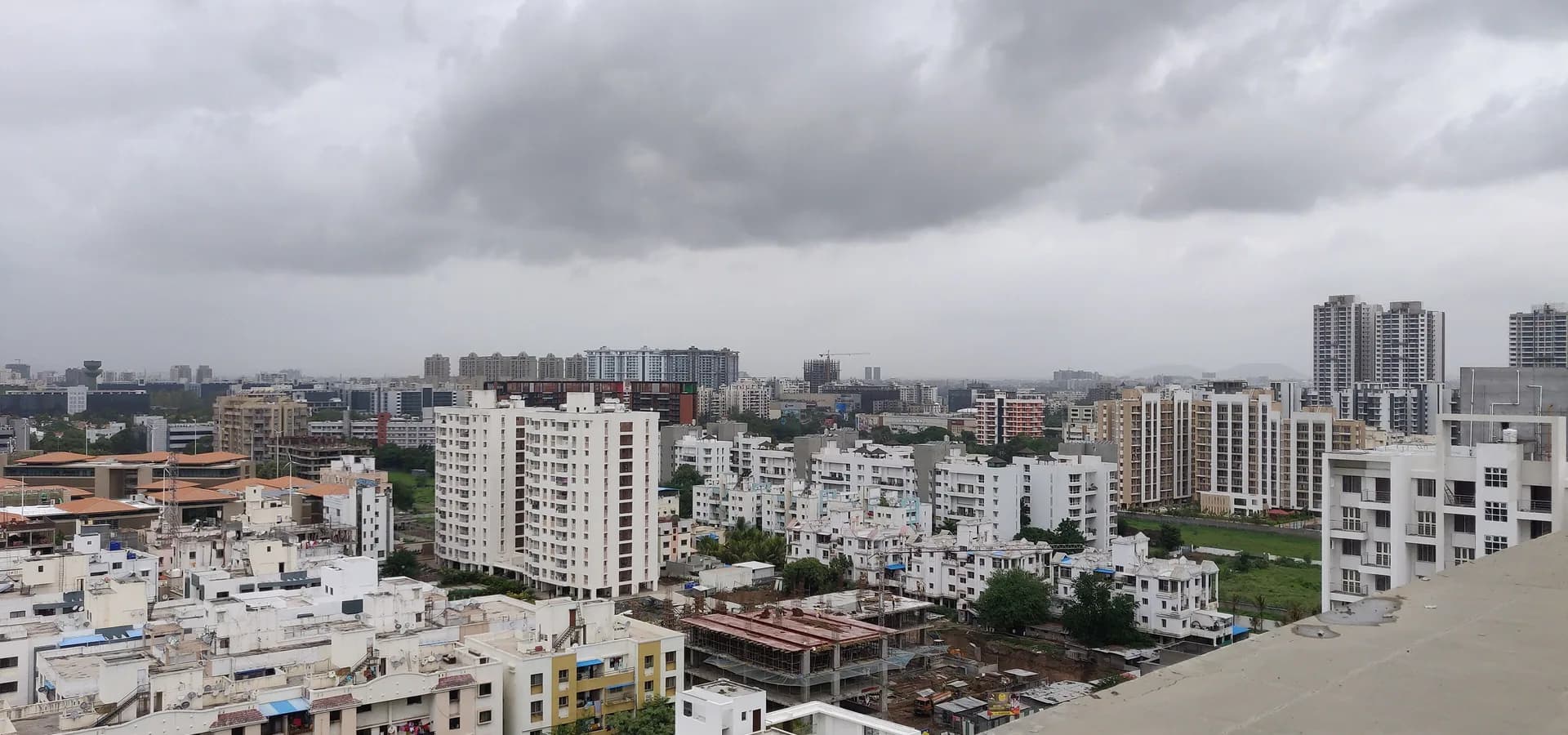 Weite Dachblick auf weiße Wohntürme und grüne Bäume in Pune unter schweren grauen Monsunwolken