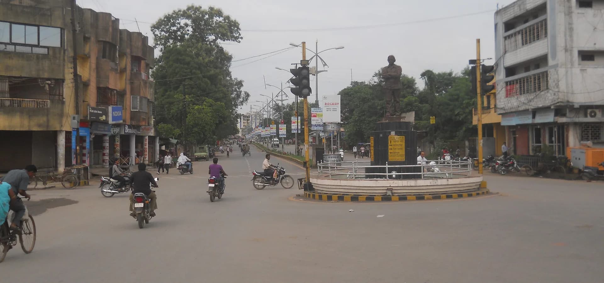 Hemu Kalani Chowk in Raigarh, ein Kreisverkehr mit Statue und Motorrädern auf einer breiten Allee mit Läden und Bäumen