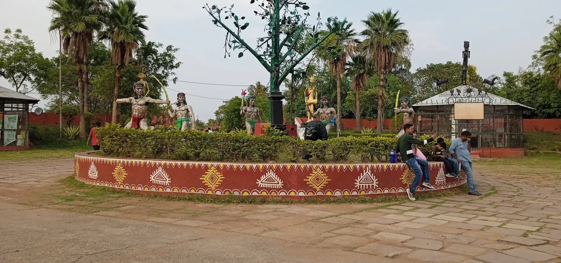 Skulpturen von Stammesjägern um einen Metallbaum auf einem bemalten Sockel, mit Palmen, in Purkhouti Muktangan, Nava Raipur