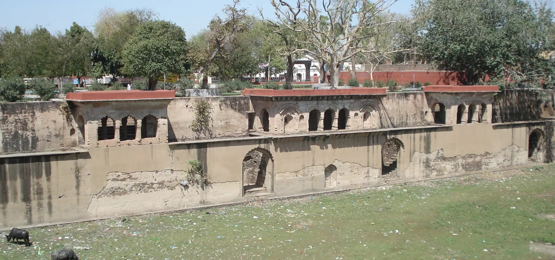 रेवाड़ी में बड़ा तालाब की पत्थर की दीवारों के साथ पुराने मेहराबदार मंडप, ऊपर पेड़ और नीला आसमान