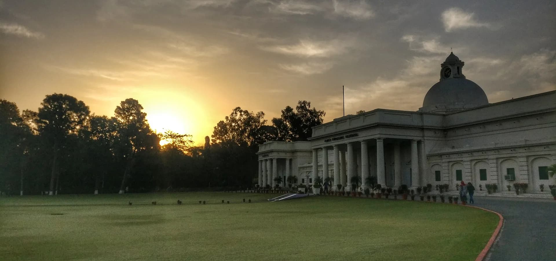 Das Hauptgebäude des IIT Roorkee mit Kuppel und weißen Säulen neben einer weiten Rasenfläche, während hinter den Bäumen die Sonne aufgeht