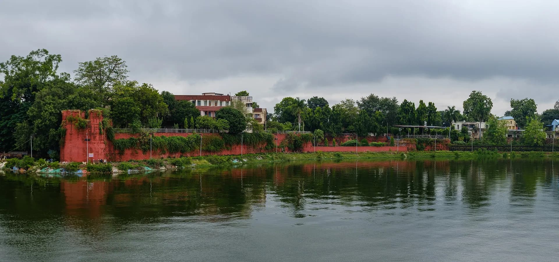Muralhas vermelhas do Forte de Sagar e templos à beira do lago refletidos na água calma do lago Lakha Banjara sob um céu nublado