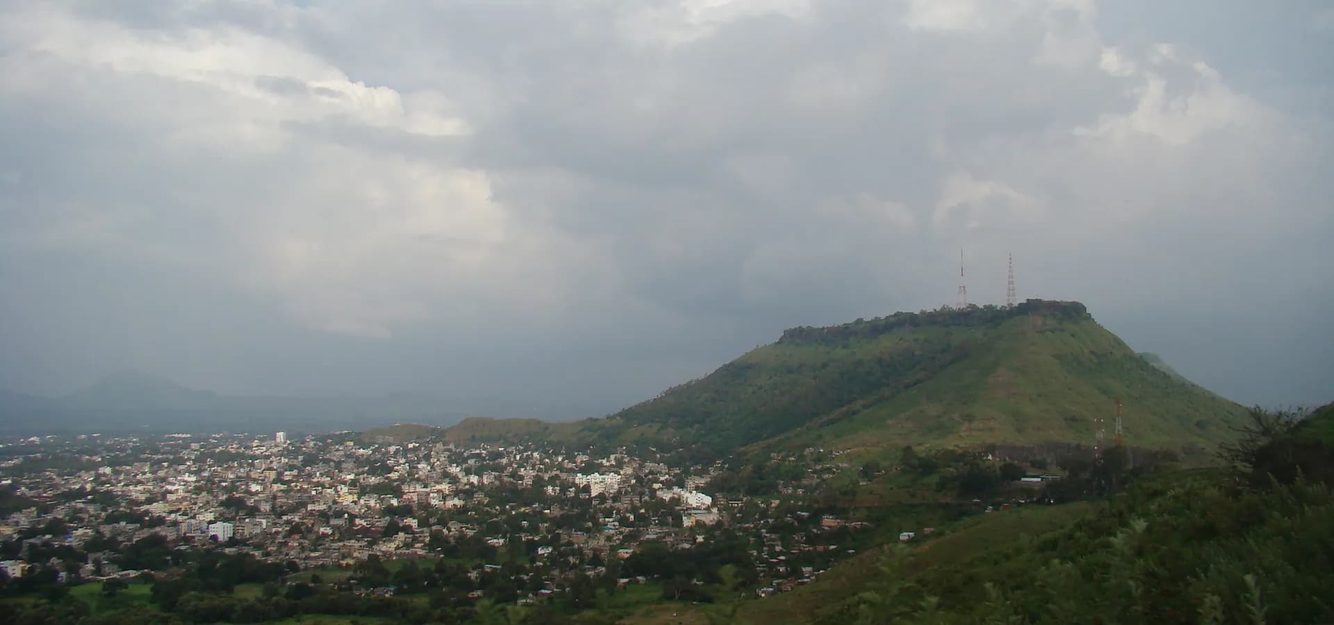 Die Stadt Satara in der Ebene unter dem grünen, flach gewölbten Hügel Ajinkyatara unter großen Monsunwolken