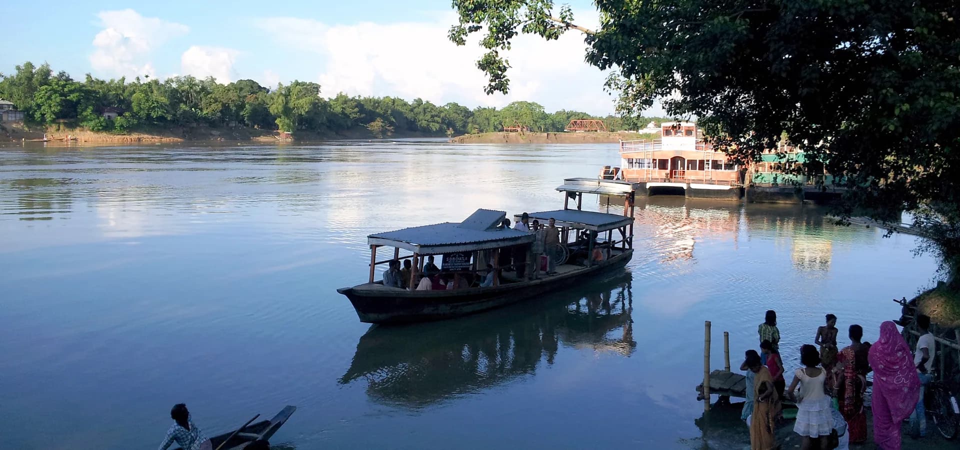 Roofed ferry crossing the calm, wide Barak River at Silchar under a blue sky with tree-lined banks