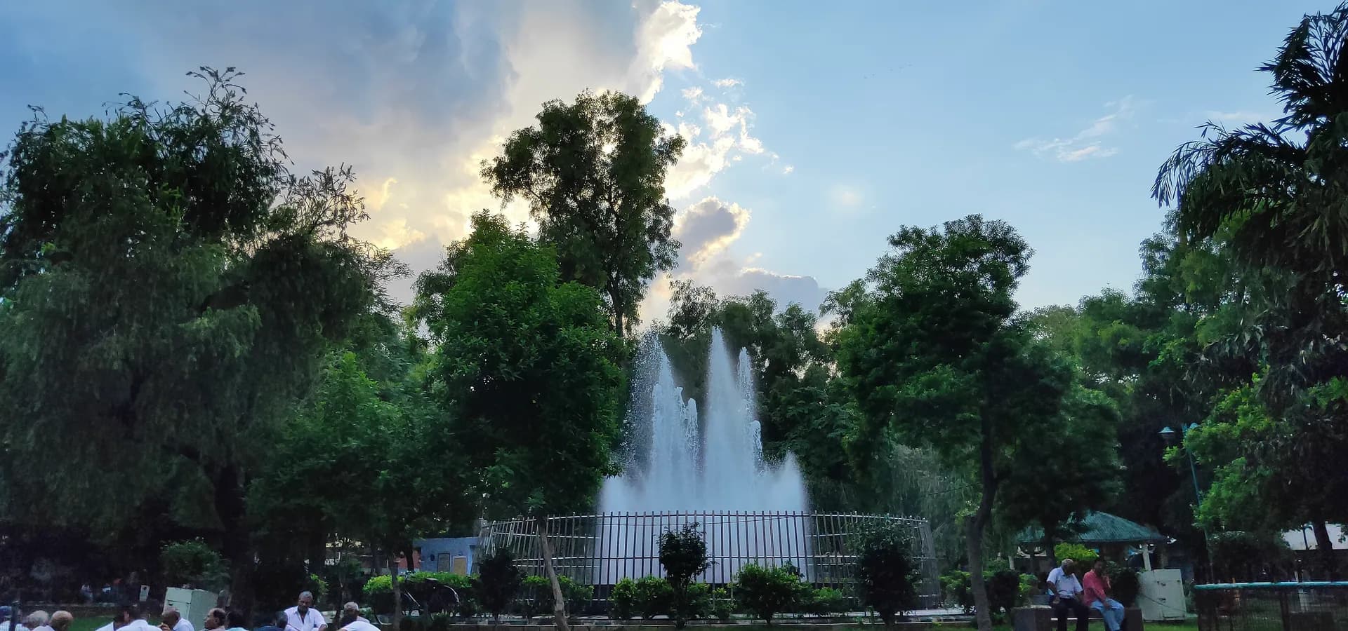 Ein hoher Springbrunnen, umrahmt von belaubten Bäumen und wolkigem Himmel, in einem Gemeinschaftspark in Sonipat