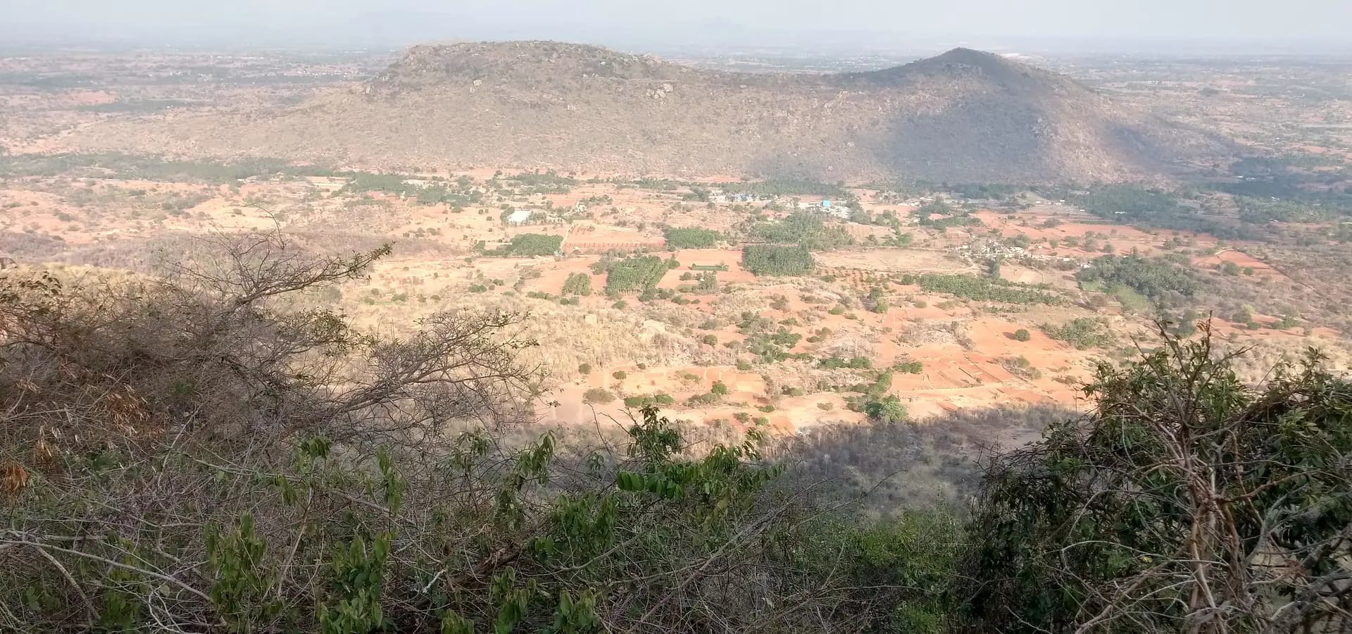 Amplia vista desde Devarayanadurga sobre campos rojizos, matorral y una colina rocosa bajo un cielo brumoso